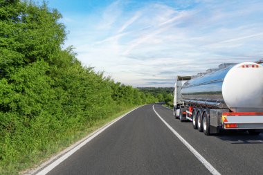 Büyük bir tanker kamyonu yeşil yemyeşil ve ince bulutlu açık mavi gökyüzü ile çevrili manzaralı kırsal bir otoyolda ilerliyor..