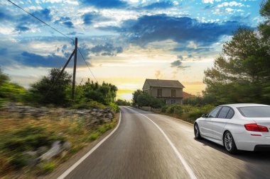 Beyaz bir araba gün batımında huzurlu bir köy evinin önünden hızla geçiyor ve kırsal alanda hareket ve özgürlük hissi yaratıyor..