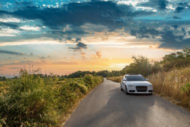 Dar bir kırsal yolda beyaz bir araba var. Etrafı yemyeşil, dramatik bir günbatımı gökyüzünün altında..