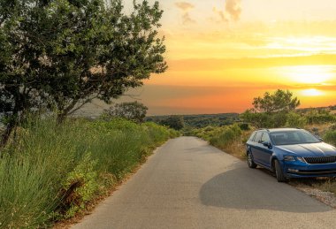Güneş yemyeşil bir arazinin üzerinde batarken, sakin bir kır yolunun yanında mavi bir araba park edilmiş, altın akşam ışığında sahneyi yıkıyor..