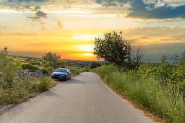 Güneş yemyeşil bir arazinin üzerinde batarken, sakin bir kır yolunun yanında mavi bir araba park edilmiş, altın akşam ışığında sahneyi yıkıyor..