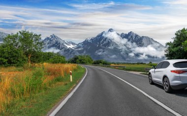 Bereketli tarlalar ve bulutların arasından yükselen dramatik karlı zirvelerle çevrili, dolambaçlı bir dağ yolunda beyaz bir araba geziniyor..