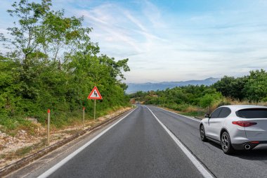 Beyaz bir araba dağlara doğru sakin kırsal bir otoyol boyunca sürüyor, sakin, mavi bir gökyüzü altında ilerleyen keskin virajlar hakkında bir trafik işaretini geçiyor..