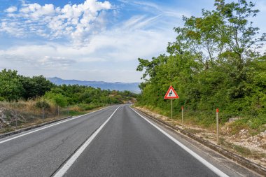Kavisli bir köy yolu, parçalı bulutlu bir gökyüzünün altındaki mesafeye kadar uzanır. Bir trafik levhası, yaklaşan kıvrımların yemyeşil yeşilliklerle çevrili olduğunu belirtir..