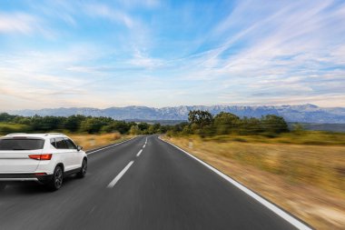 Beyaz bir SUV, canlı mavi gökyüzünün altında kırsal ve dağlarla çevrili manzaralı, açık bir yolda hızla ilerliyor, özgürlüğün ve maceranın özünü yakalıyor..