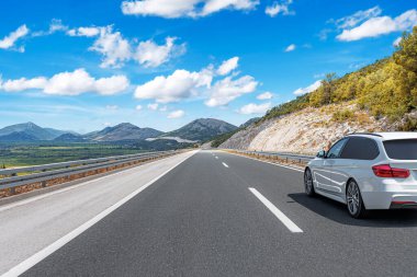 Parlak beyaz bir araba, dağlarla çevrili geniş ve boş bir otoyol boyunca ilerliyor..