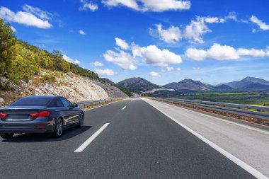 Parlak mavi bir gökyüzünün altında dağınık bulutlarla manzaralı bir dağ yolunda giden parlak siyah bir araba..
