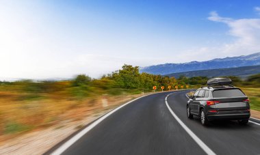 Çatı kargo kutusu olan modern bir SUV, açık mavi gökyüzünün altında ağaçlar ve dağlarla çevrili dolambaçlı bir köy yolunda ilerliyor..
