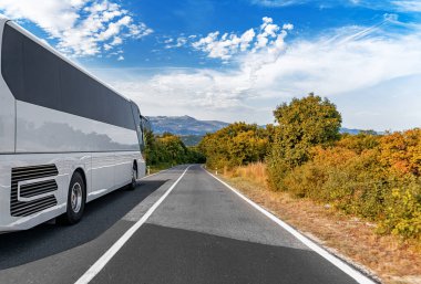 Beyaz bir tur otobüsü, sonbahar ağaçlarıyla çevrili manzaralı kırsal bir yolda, dağınık bulutlarla dolu canlı mavi bir gökyüzü altında seyahat ediyor..