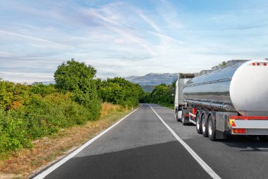 Gri bir tanker, yemyeşil ve sakin mavi gökyüzünün altındaki uzak dağlarla çevrili sakin bir kırsal yolda ilerliyor..