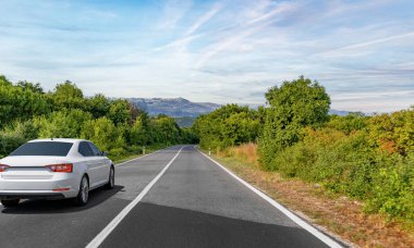 Beyaz bir sedan, berrak mavi gökyüzünün altında yemyeşil ve uzak dağlarla çevrili huzurlu bir kırsal yolda ilerliyor..