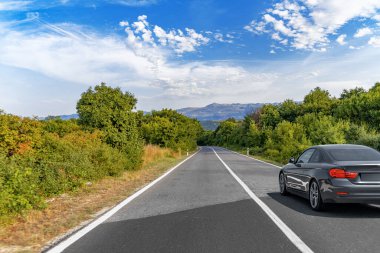 Pürüzsüz gri bir araba, parlak mavi bir gökyüzünün altında yeşil ve dağlarla kaplı sakin bir köy yolunda ilerliyor..