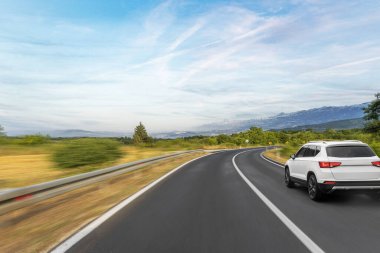 Beyaz bir SUV, yeşil ağaçlar ve bulutlu bir gökyüzü altında dağ manzarası ile çevrili keskin dönüş işaretleri ile virajlı kırsal bir yol boyunca sürer..