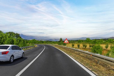 Beyaz bir sedan, metal bariyerler ve çarpma uyarısı ile dolambaçlı bir kırsal yol boyunca ilerliyor. Etrafı tarlalar, ağaçlar ve açık gökyüzünün altındaki uzak dağlarla çevrili..