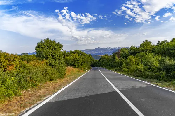 Sakin bir çift şeritli yol, yeşil ağaçlar ve dağlarla dolu verimli kırsal bölgelere doğru uzanır. Dalgalı mavi gökyüzü altında dağınık bulutlar..