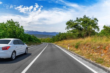 Beyaz bir sedan, yemyeşil yemyeşil ve dağlarla çevrili dolambaçlı kırsal bir yol boyunca ilerliyor. Parlak mavi bir gökyüzünün altında dağınık bulutlarla..