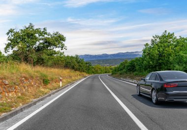 Pürüzsüz siyah bir araba dolambaçlı kırsal bir yolda ilerliyor. Etrafı yemyeşil ve açık mavi gökyüzünün altındaki uzak dağlarla çevrili..