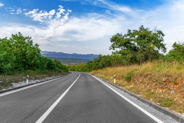 Dolambaçlı asfalt bir yol yeşil kırsal bir arazide, dağınık bulutlar ve uzak dağlarla dolu canlı mavi bir gökyüzü altında yavaşça kıvrılır..