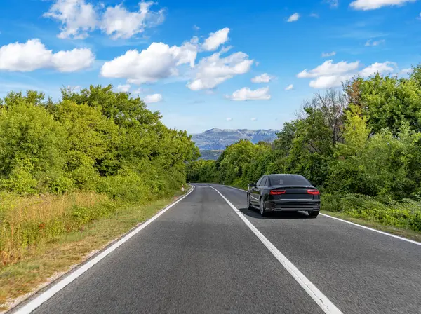 Pürüzsüz, manzaralı bir yolda ilerleyen siyah lüks bir sedan yoğun yeşillik ve dağ manzarasıyla kaplı tüylü beyaz bulutlu, parlak mavi bir gökyüzünün altında..