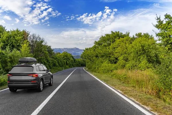 Çatı kutusuna sahip bir SUV, bereketli yeşil kırlarda, bulutların ve dağılmış dağların altında dolambaçlı, dolambaçlı bir yolda ilerliyor..