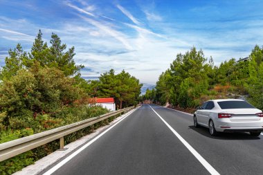 Beyaz bir sedan, bulutlarla kaplı parlak bir gökyüzünün altında manzaralı huzurlu bir orman yolunda ilerliyor..
