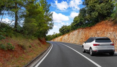 Beyaz bir SUV, dolambaçlı bir orman yolunda, parlak mavi bir gökyüzünün altında, dağınık bulutlarla ilerliyor..