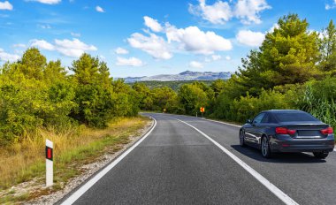 Parlak mavi gökyüzünün altında, dağınık beyaz bulutlarla kıvrımlı bir orman yolunda ilerleyen parlak siyah bir coupe..