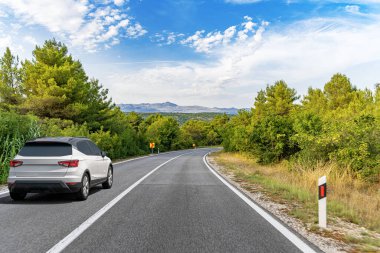 Beyaz bir SUV, dolambaçlı bir orman yolunda, parlak mavi bir gökyüzünün altında, dağınık bulutlarla ilerliyor..