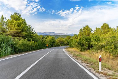Dolambaçlı asfalt bir yol yeşil kırsal bir arazide, dağınık bulutlar ve uzak dağlarla dolu canlı mavi bir gökyüzü altında yavaşça kıvrılır..