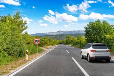Beyaz bir SUV, dolambaçlı bir orman yolunda, parlak mavi bir gökyüzünün altında, dağınık bulutlarla ilerliyor..