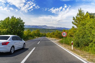 Beyaz bir sedan, yemyeşil yemyeşil ve dağlarla çevrili dolambaçlı kırsal bir yol boyunca ilerliyor. Parlak mavi bir gökyüzünün altında dağınık bulutlarla..