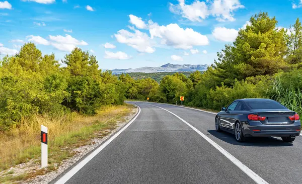 Parlak mavi gökyüzünün altında, dağınık beyaz bulutlarla kıvrımlı bir orman yolunda ilerleyen parlak siyah bir coupe..