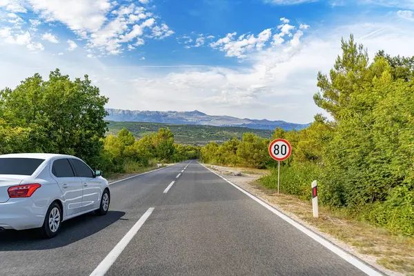 Beyaz bir sedan, yemyeşil yemyeşil ve dağlarla çevrili dolambaçlı kırsal bir yol boyunca ilerliyor. Parlak mavi bir gökyüzünün altında dağınık bulutlarla..