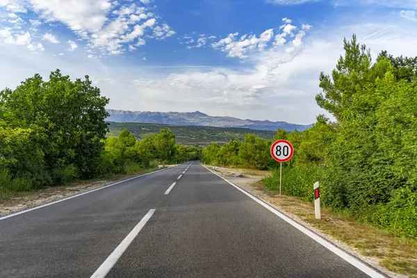 Dolambaçlı asfalt bir yol yeşil kırsal bir arazide, dağınık bulutlar ve uzak dağlarla dolu canlı mavi bir gökyüzü altında yavaşça kıvrılır..