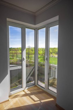 Modern bir iç mekânda, güneşli bir köşe penceresi berrak mavi gökyüzünün altında sakin yeşil bir manzarayı gözler önüne seriyor..