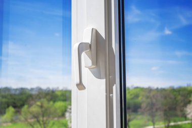 Beyaz plastik bir pencere kolunun yakın çekimi arka planda parlak bir bahar manzarası olan temiz modern tasarımı gösterir..