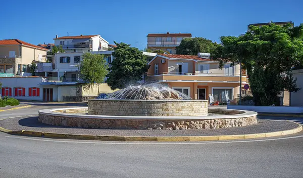 Primosten 'de açık mavi bir gökyüzünün altında, Akdeniz tarzı evlerle çevrili sakin bir kavşağın ortasına su püskürten dairesel taş çeşme..