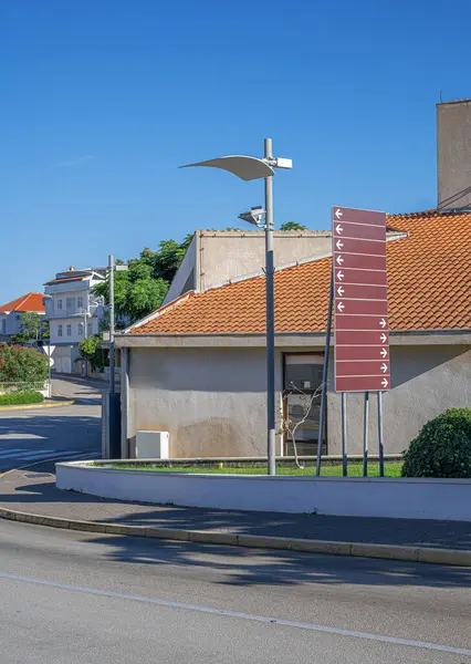 Modern sokak lambasıyla sokak manzarası, çoklu oklu bir yön işareti ve açık mavi gökyüzünün altında kırmızı kiremitli çatıları olan binalar..