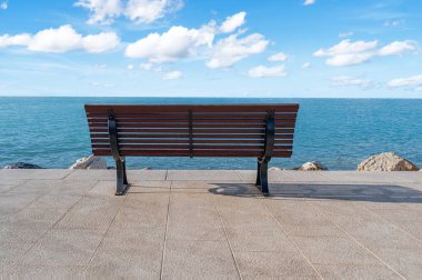 Deniz kenarındaki boş bir bankta, sakin okyanusa bakan açık mavi gökyüzünün altında dağınık beyaz bulutlar, huzur ve yalnızlık hissi uyandırıyor..