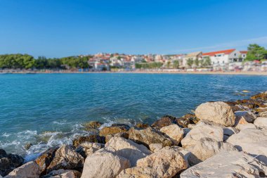 Güneşli çakıl taşlı plaj, kıyıya vuran turkuaz dalgalar, sazlı şemsiyelerin altında dinlenen güneş banyosu, ve arka planda bir kıyı köyü..