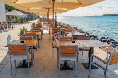 Huzurlu deniz kenarı restoranı, modern ahşap masalar ve büyük şemsiyelerin altında deniz manzaralı bir gezinti alanı boyunca sıralanmış sandalyeler..