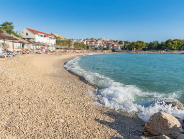 Güneşli çakıl taşlı plaj, kıyıya vuran turkuaz dalgalar, sazlı şemsiyelerin altında dinlenen güneş banyosu, ve arka planda bir kıyı köyü..