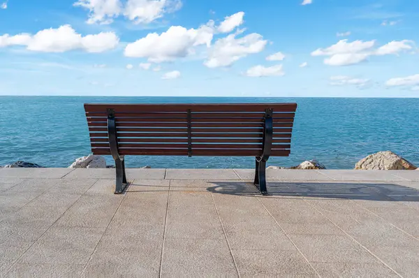Deniz kenarındaki boş bir bankta, sakin okyanusa bakan açık mavi gökyüzünün altında dağınık beyaz bulutlar, huzur ve yalnızlık hissi uyandırıyor..
