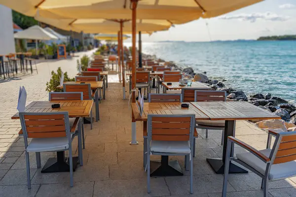Huzurlu deniz kenarı restoranı, modern ahşap masalar ve büyük şemsiyelerin altında deniz manzaralı bir gezinti alanı boyunca sıralanmış sandalyeler..