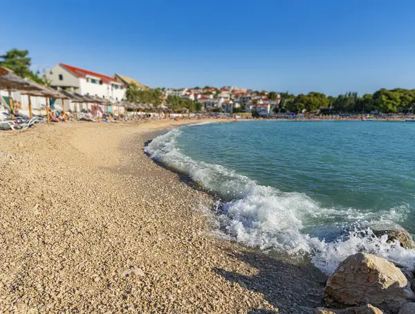 Güneşli çakıl taşlı plaj, kıyıya vuran turkuaz dalgalar, sazlı şemsiyelerin altında dinlenen güneş banyosu, ve arka planda bir kıyı köyü..