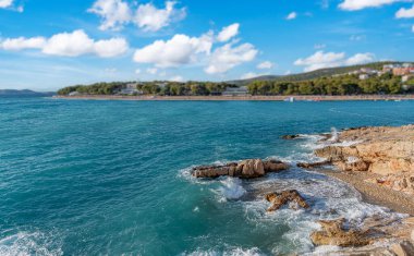 Güçlü Adriyatik dalgaları, ormanlık bir kıyı şeridi ve açık bir gökyüzünün altında görünen sahil binalarıyla kayalık kıyılara çarpıyor..