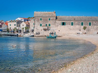 Hırvatistan 'ın Primosten kentindeki sakin bir çakıl taşı plajı boyunca açık mavi bir gökyüzünün altında yeşil kepenkleri olan taştan bir bina duruyor..