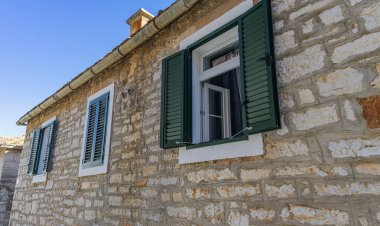 Güneşli bir günde açık mavi gökyüzünün altında yeşil kepenkleri ve açık pencereleri olan büyüleyici taştan bir ev..