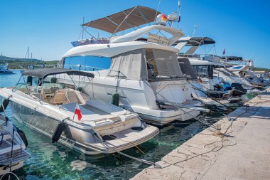 Birçok lüks yat ve tekne, sakin deniz ve uzak yeşil kıyı şeridi ile çevrili güneşli bir marinada demirlemiştir.