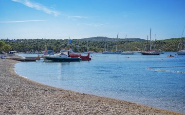 Hırvatistan 'ın Primosten kentinde parlak mavi bir gökyüzünün altında berrak turkuaz sular, demirlemiş yatlar ve yeşil bir yamaçtan oluşan sakin bir kıyı manzarası.